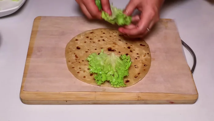 Assemble the roll: Place a paratha on a butter paper. Spread some salad leaves, cooked chicken, onion, and cheese sauce on the paratha. Roll it tightly.