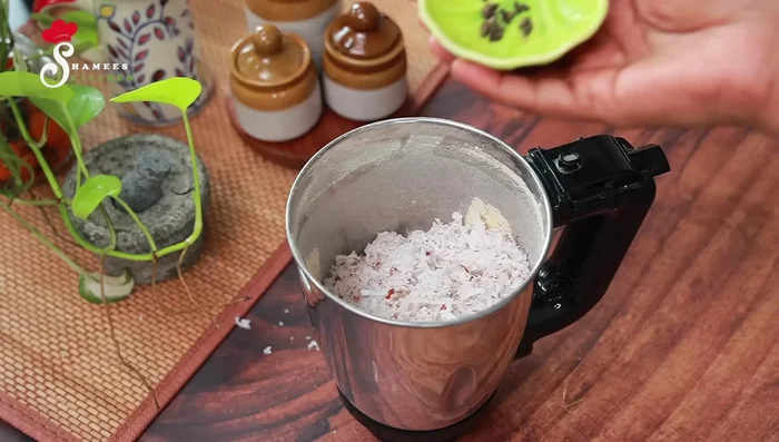 Grate a tablespoon of coconut and add it to the mixer along with the ground chickpeas, cardamom seeds, and half a cup of water. Grind to a coarse consistency.